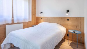 A bedroom with a wooden headboard, a bed with white bedding, a small round table with a remote, and two wall-mounted lights.