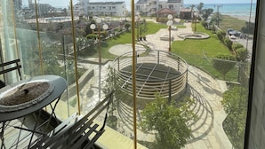 A balcony with a view of a landscaped area, a circular water feature, and buildings in the distance. - Duplex at Solemar Resort, Totally Renovated, Pools, Tennis, Basketball, Football (Jounieh)
