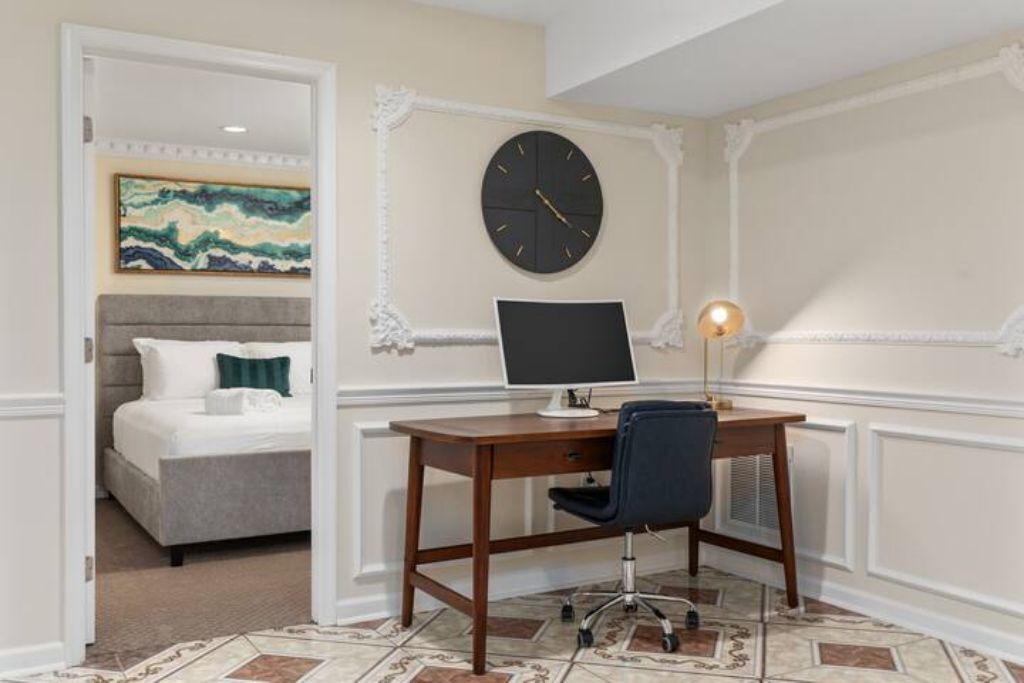 Een hotelkamer met een bed, een bureau met een computer, een klok en een schilderij aan de muur.