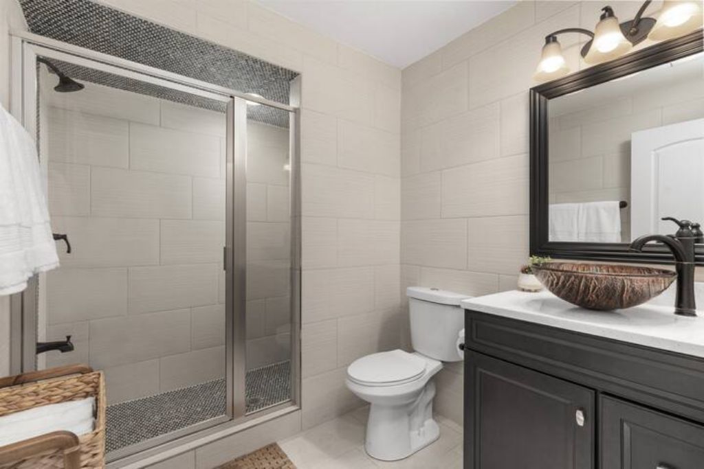 Un baño con ducha de vidrio, inodoro blanco, tocador oscuro con un espejo grande y un lavabo.