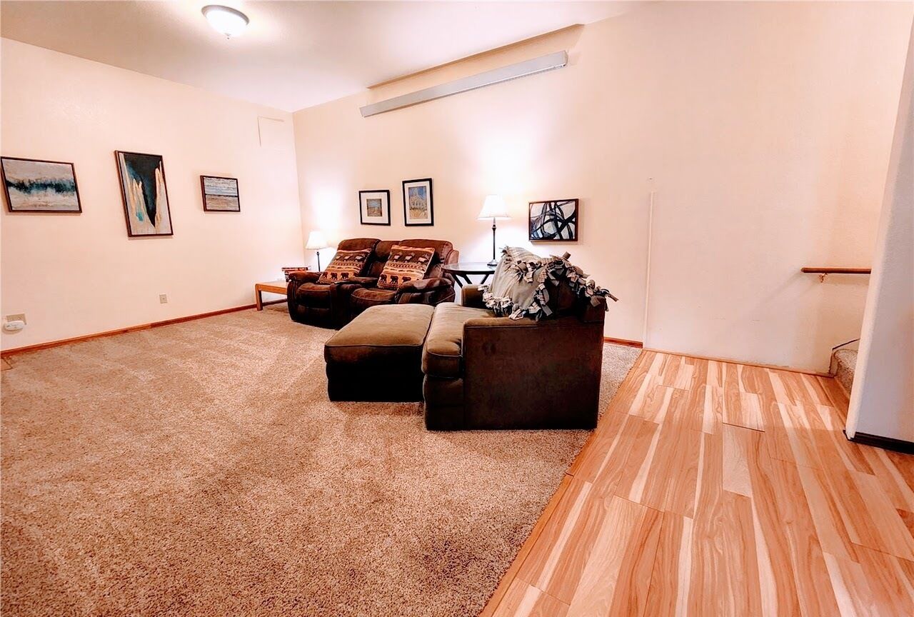 A spacious living room with a large brown carpet, a brown sofa, and a wooden floor. There are framed pictures on the walls.