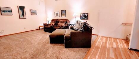 A spacious living room with a large brown carpet, a brown sofa, and a wooden floor. There are framed pictures on the walls.
