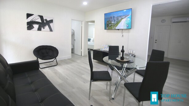 A modern living room with a black sofa, a glass dining table with chairs, a flat-screen TV on the wall, and a door leading to another room. - Diaz Stay Miami Airport (Miami)