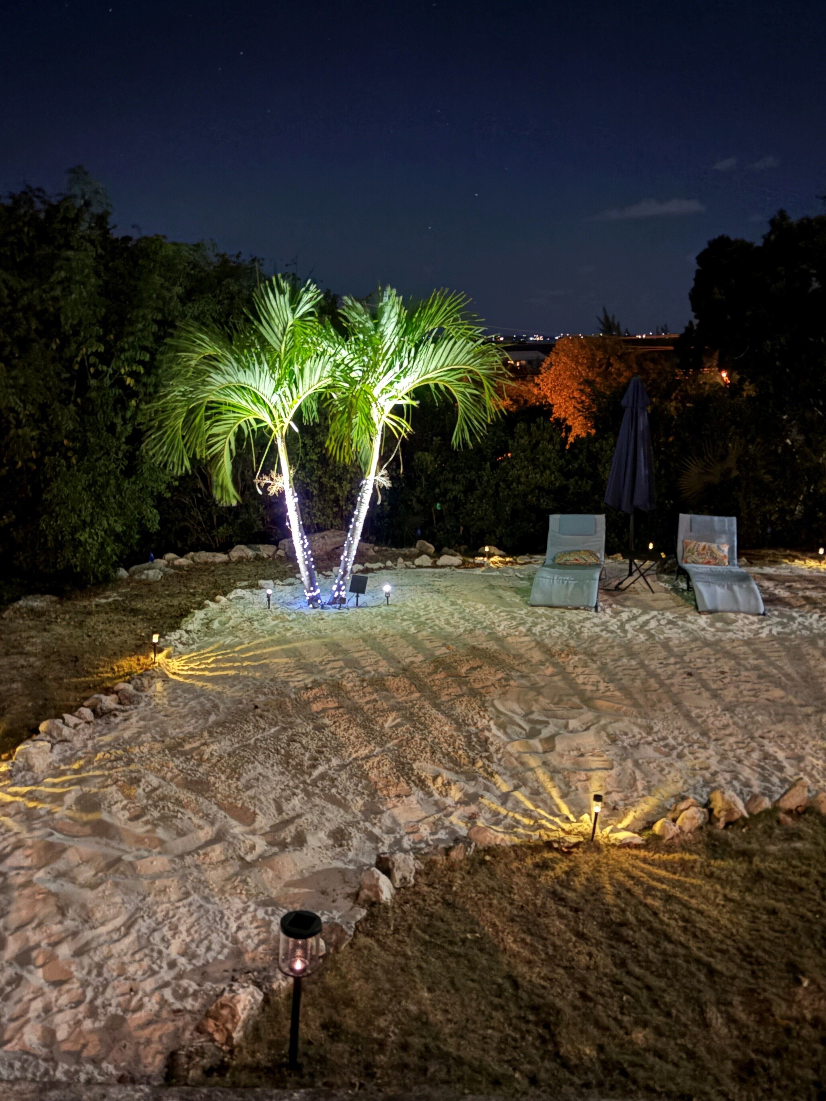 A nighttime outdoor area with a palm tree illuminated by lights, a seating area with chairs and an umbrella, and a sandy ground.