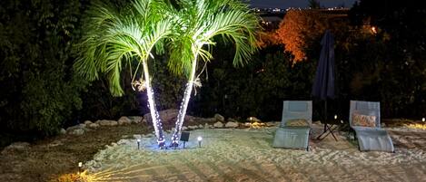A nighttime outdoor area with a palm tree illuminated by lights, a seating area with chairs and an umbrella, and a sandy ground.