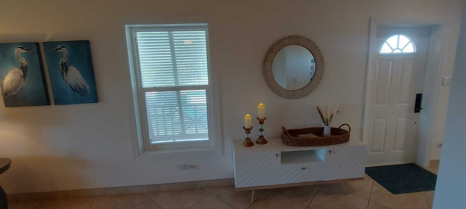 A hallway with a white door, a window with blinds, a mirror, and two wall-mounted paintings.