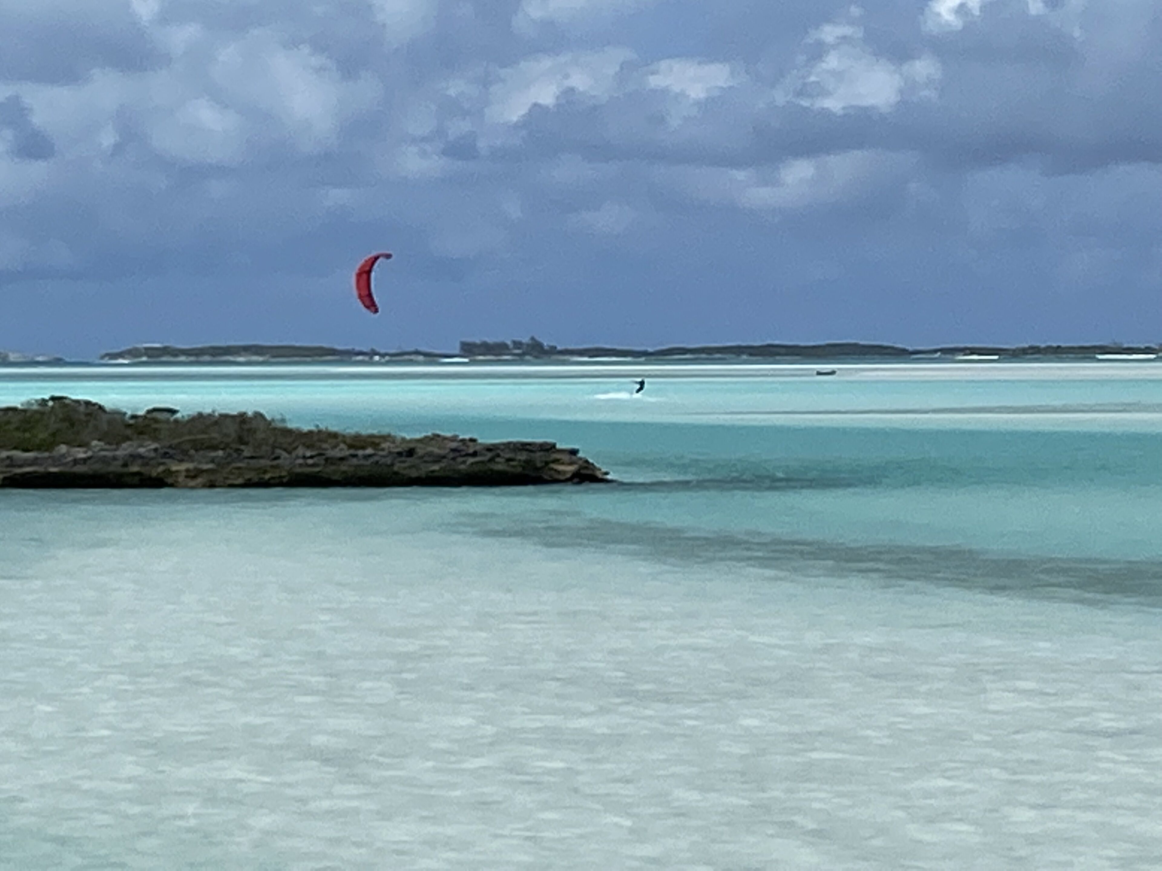 Una playa con agua turquesa cristalina, una costa rocosa y una persona practicando kitesurf a lo lejos.