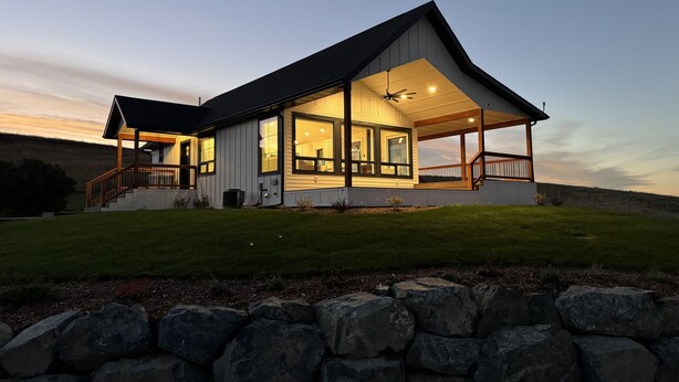 A modern house with a large porch, a staircase, and a stone wall in the foreground. - Modern Farmhouse with Breathtaking Views! (Bozeman)