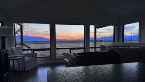 A modern living room with a large window offering a view of a sunset over mountains. - Modern Farmhouse with Breathtaking Views! (Bozeman)