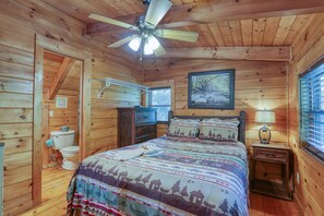 Ein Schlafzimmer einer Holzhütte mit Bett, Nachttisch, Deckenventilator, Fenster mit Jalousien und einem gerahmten Bild an der Wand.