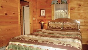 Een slaapkamer in een houten hutje, met een bed, nachtkastje, lamp en een raam met luiken.