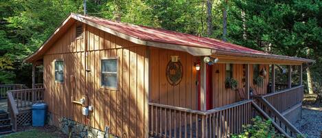 A wooden cabin with a porch, stone foundation, and a red roof.