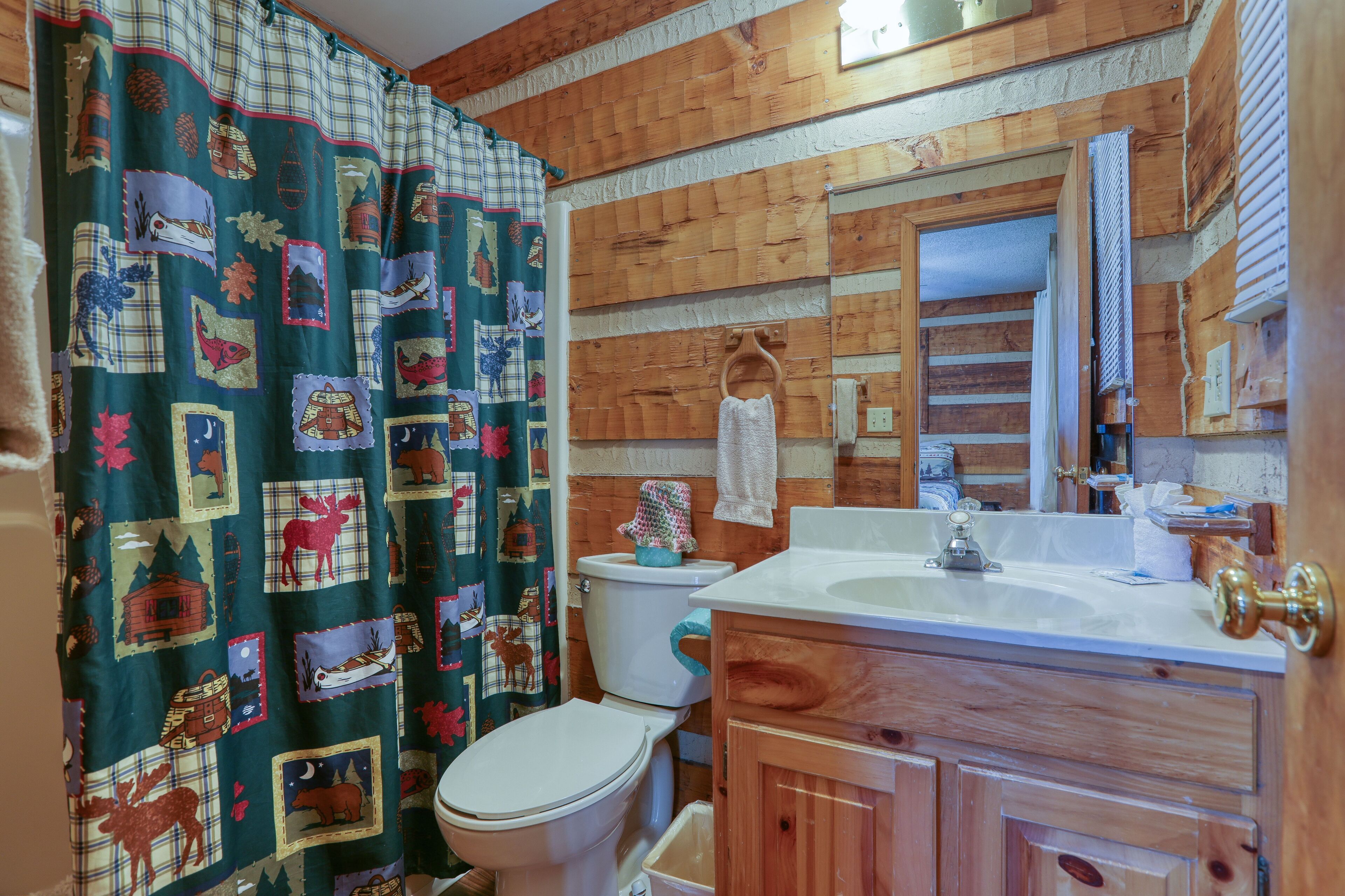 A bathroom with a wooden wall, a white toilet, a sink with a mirror, and a shower curtain with a forest and animal pattern.