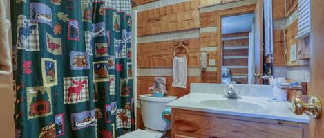 A bathroom with a wooden wall, a white toilet, a sink with a mirror, and a shower curtain with a forest and animal pattern.