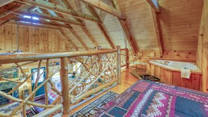 A wooden cabin interior with a loft bedroom, a stone fireplace, and a hot tub.