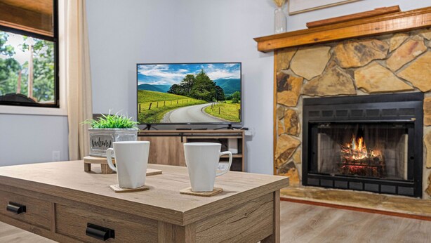 Een houten tafel met twee witte mokken, een televisiescherm waarop een schilderachtig landschap te zien is, en een stenen open haard.