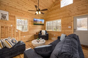 Interior acolhedor de uma cabana, com decoração em madeira, ventilador de teto, sofá, mesa de centro e porta com janelas.