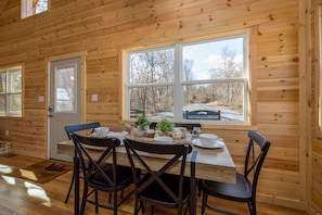 A wooden cabin interior with a dining table set for four, large windows, and a view of the outdoors.