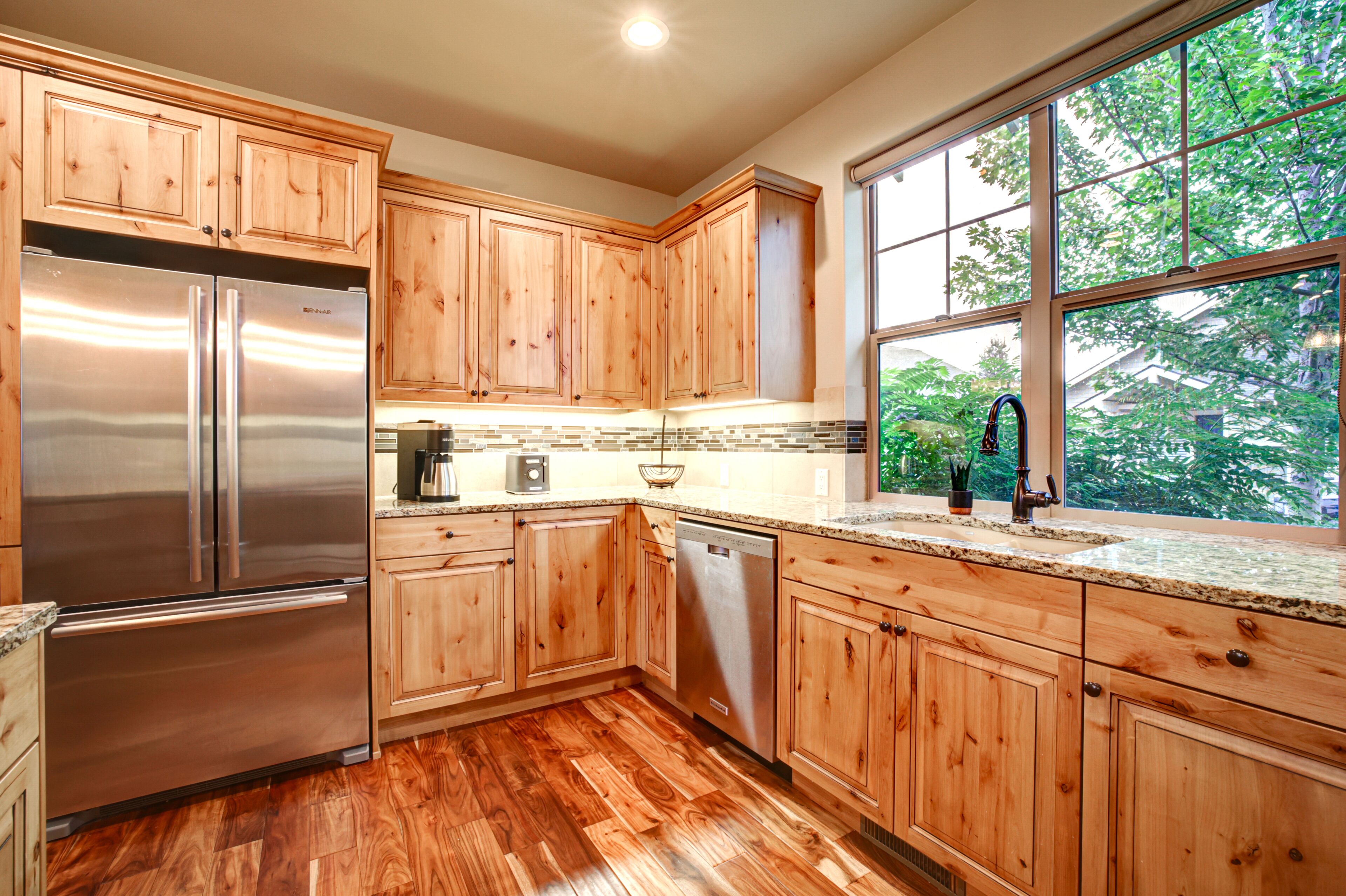 A kitchen with wooden cabinets, stainless steel appliances, a granite countertop, and a window with a view of trees.