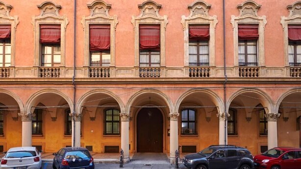 A building with a central entrance, multiple windows with red curtains, and a series of arches. - Palazzo di Alcina-Residenza D'Epoca (Bologna)