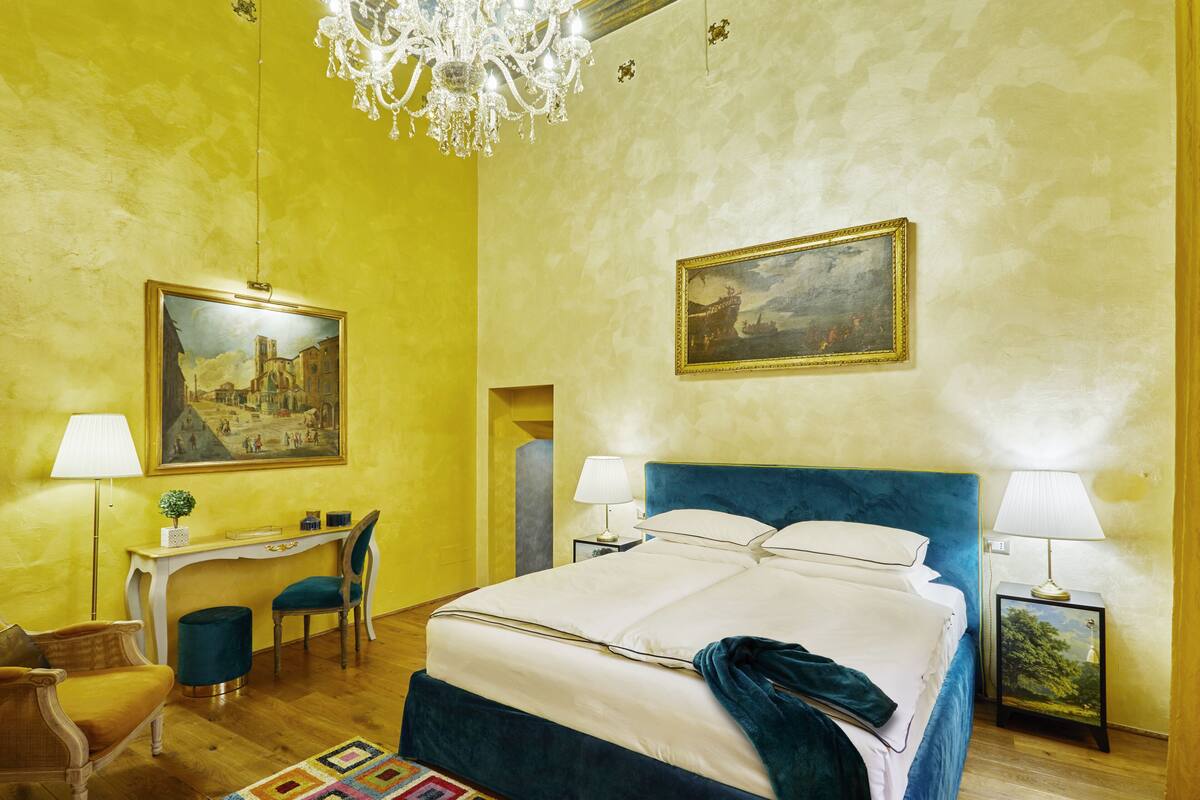 A hotel room with a large bed, two bedside tables with lamps, a desk with a chair, a chandelier, and two paintings on the walls.