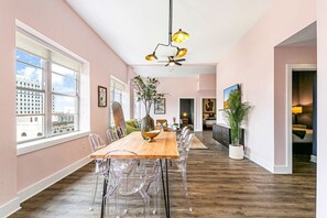 Un comedor amplio con una larga mesa de madera y sillas transparentes, ventanas grandes que ofrecen vistas a edificios y un ventilador de techo con múltiples lámparas.