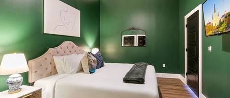 A bedroom with a bed, a nightstand, a lamp, a mirror, and a wall-mounted TV.