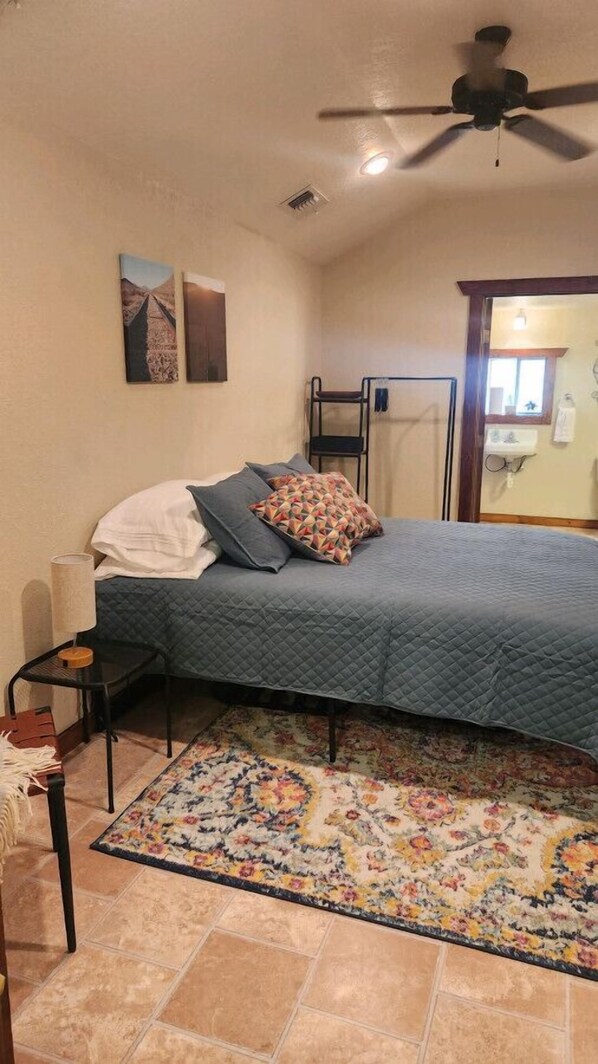 Un dormitorio con una cama, una mesita de noche, una silla, un ventilador de techo y una ventana.