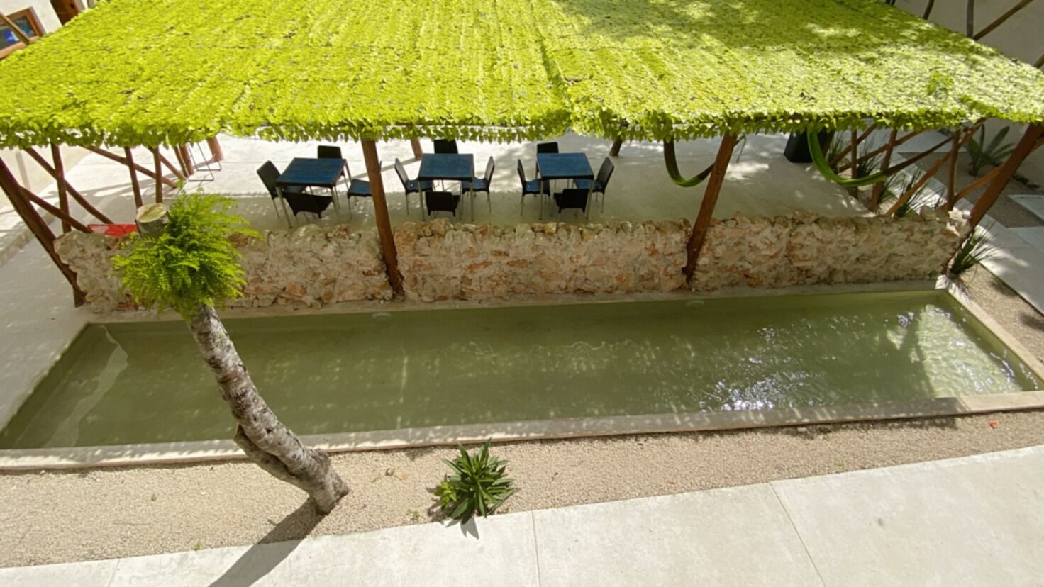 Un área de estar al aire libre con mesas y sillas bajo un dosel verde, una pequeña fuente de agua y un muro de piedra.