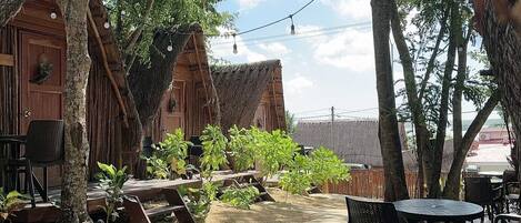 Un área de estar al aire libre rústica, con mesas y sillas de madera, rodeada de árboles y un edificio con techo de paja.