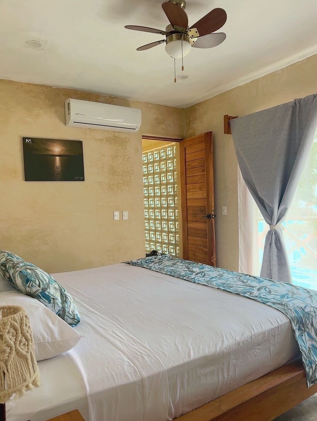 A bedroom with a bed, a ceiling fan, an air conditioning unit, a window with curtains, and a wooden door.