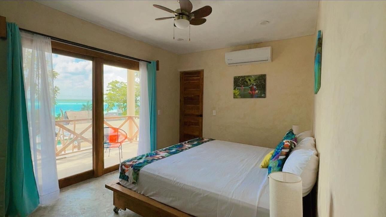 A bedroom with a bed, a ceiling fan, a window with curtains, and a view of the ocean.