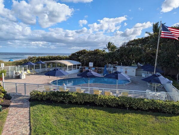A pool area with umbrellas, a flag, and a view of the ocean. - Berkshire on the Ocean (Delray Beach)