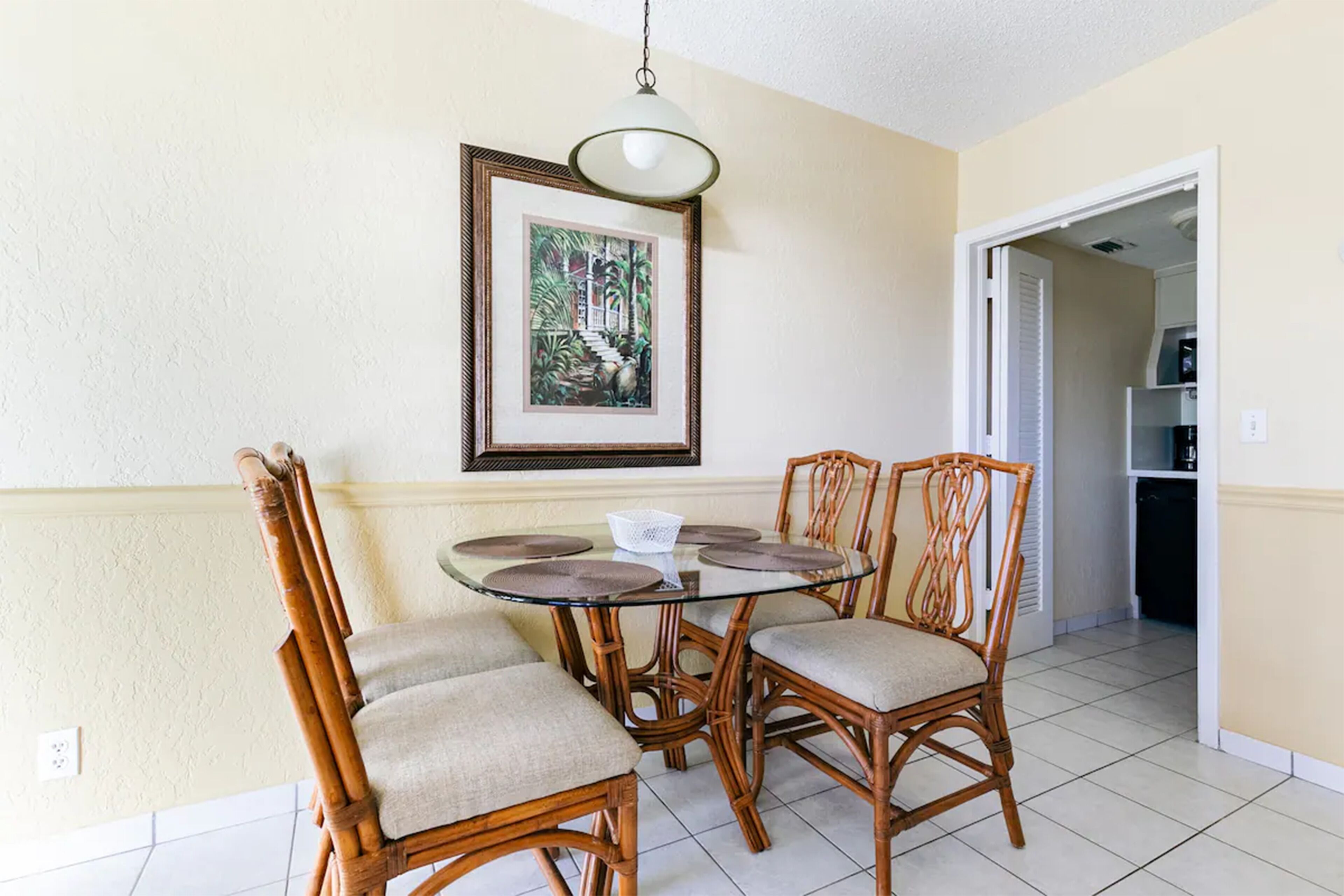 A dining area with a round glass table and four wicker chairs, a framed picture on the wall, and a kitchen visible through an open doorway.