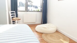 A bedroom with a bed, a chair with a cushion, a round white pouf, a woven rug, and a window with curtains.