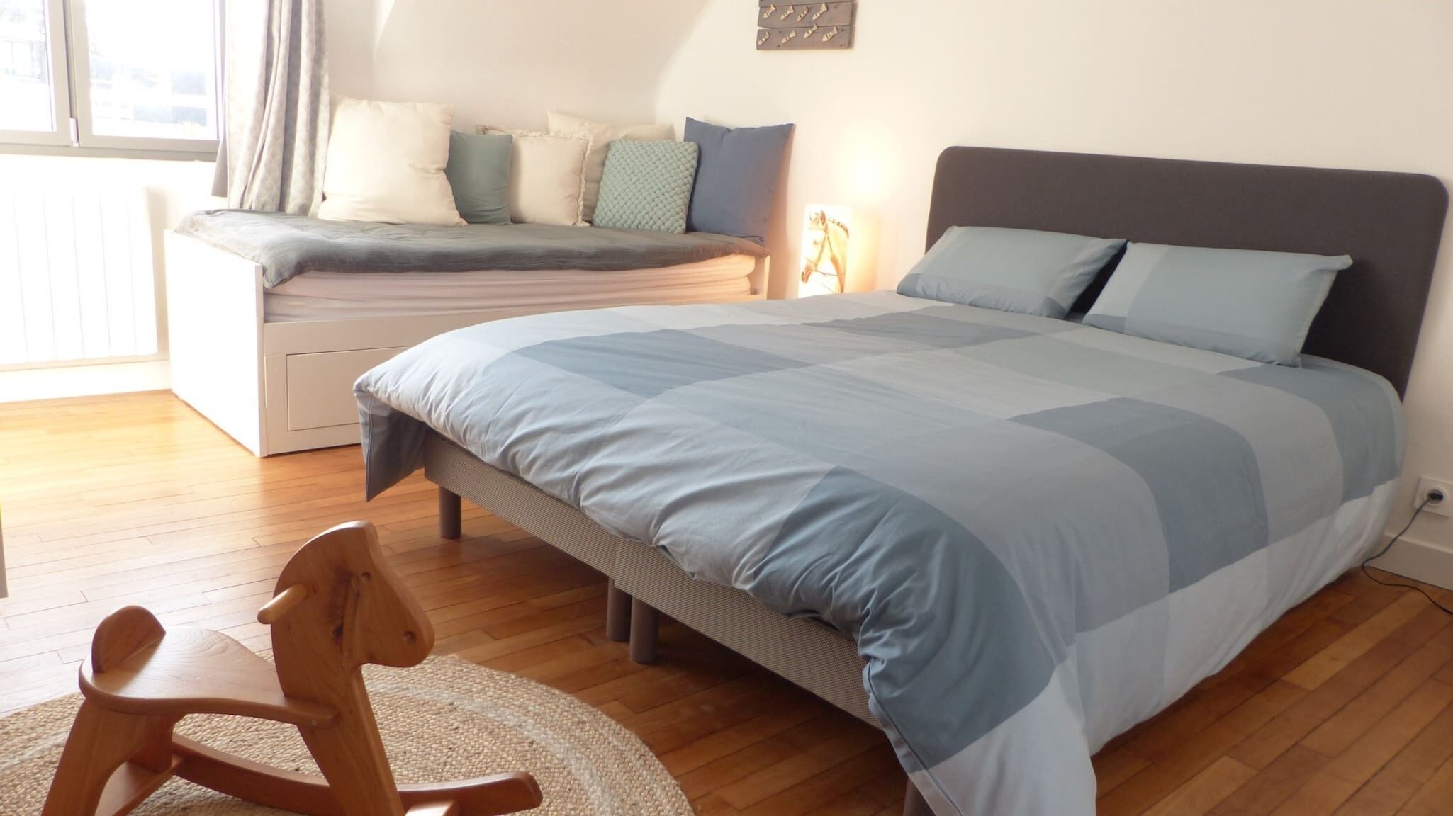 Une chambre à coucher avec un lit, un banc, un fauteuil à bascule et une fenêtre avec des rideaux.