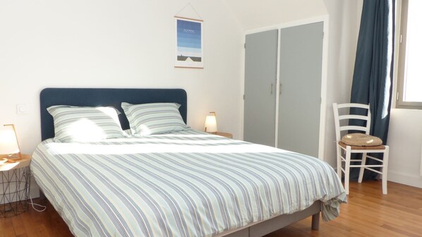 A neatly made bed with striped bedding, a bedside lamp, a chair with a cushion, and a wardrobe.