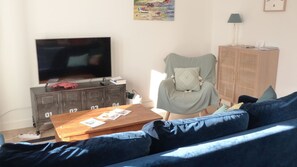 A living room with a TV, a wooden coffee table with magazines, a blue sofa, a chair, and a cabinet.
