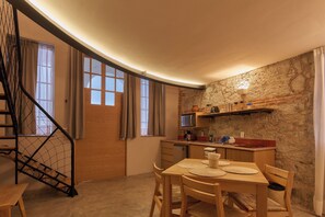 Uma cozinha compacta com uma mesa de madeira, cadeiras e micro-ondas. Uma escada com guarda-mão de metal é visível à esquerda.