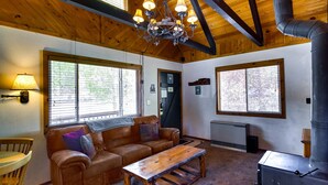 Ein gemütliches Wohnzimmer mit Ledersofa, Holztisch und einer Kristallleuchter. Große Fenster sorgen für natürliches Licht und bieten einen Ausblick nach draußen.
