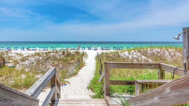 A wooden boardwalk leading to a beach with clear blue water and umbrellas.