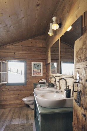 Ein Badezimmer einer Holzhütte mit einem großen Spiegel, zwei Waschbecken und einem Fenster mit Blick auf eine verschneite Landschaft.