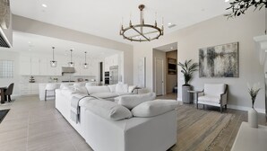 Een ruime woonkamer met een grote witte bank, een kroonluchter en een minimalistische keuken op de achtergrond.