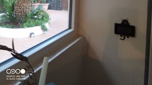 Ein Zimmer mit Fensterblick auf einen Garten, einer Wanduhr und einem kleinen schwarzen Gegenstand an der Wand.