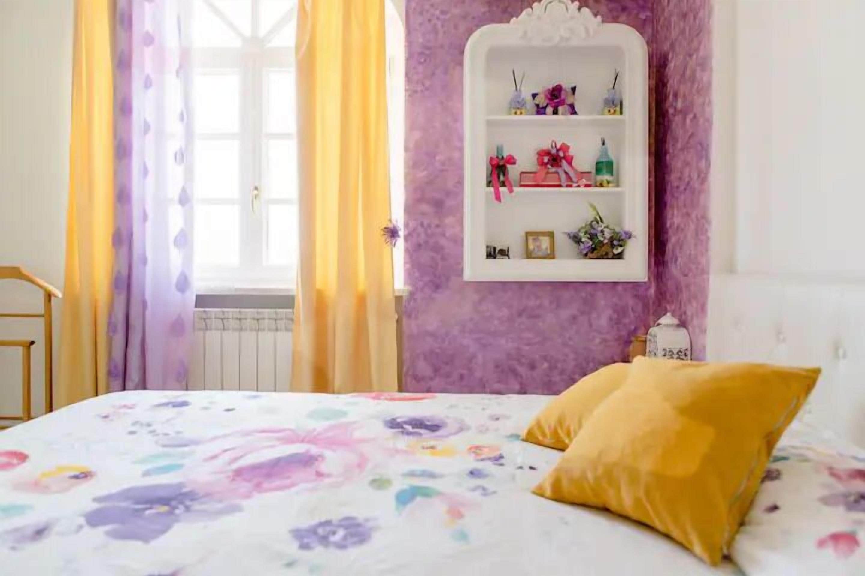Un dormitorio con edredón estampado con motivos florales, almohada amarilla, estante blanco con objetos decorativos y ventana con cortinas amarillas.