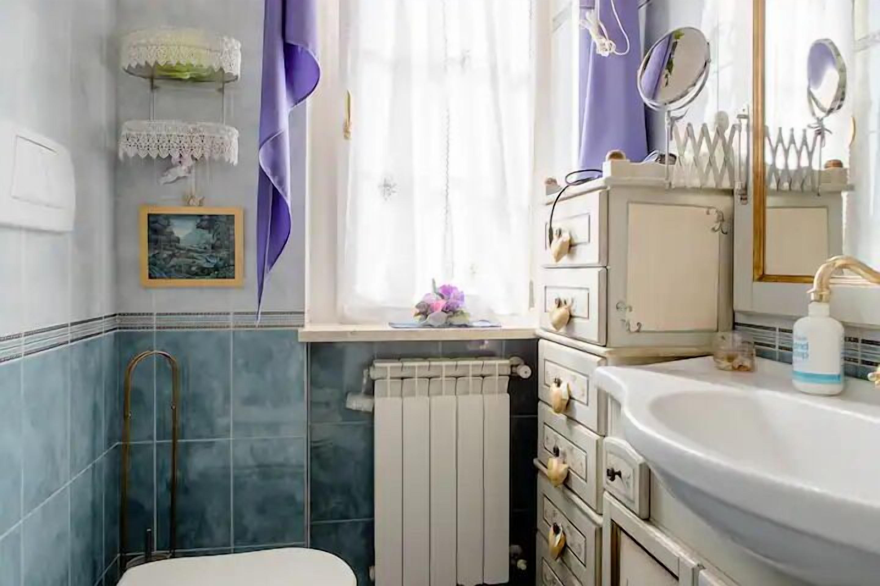 Une salle de bain avec un lavabo blanc, un miroir, un radiateur et une fenêtre avec des rideaux.