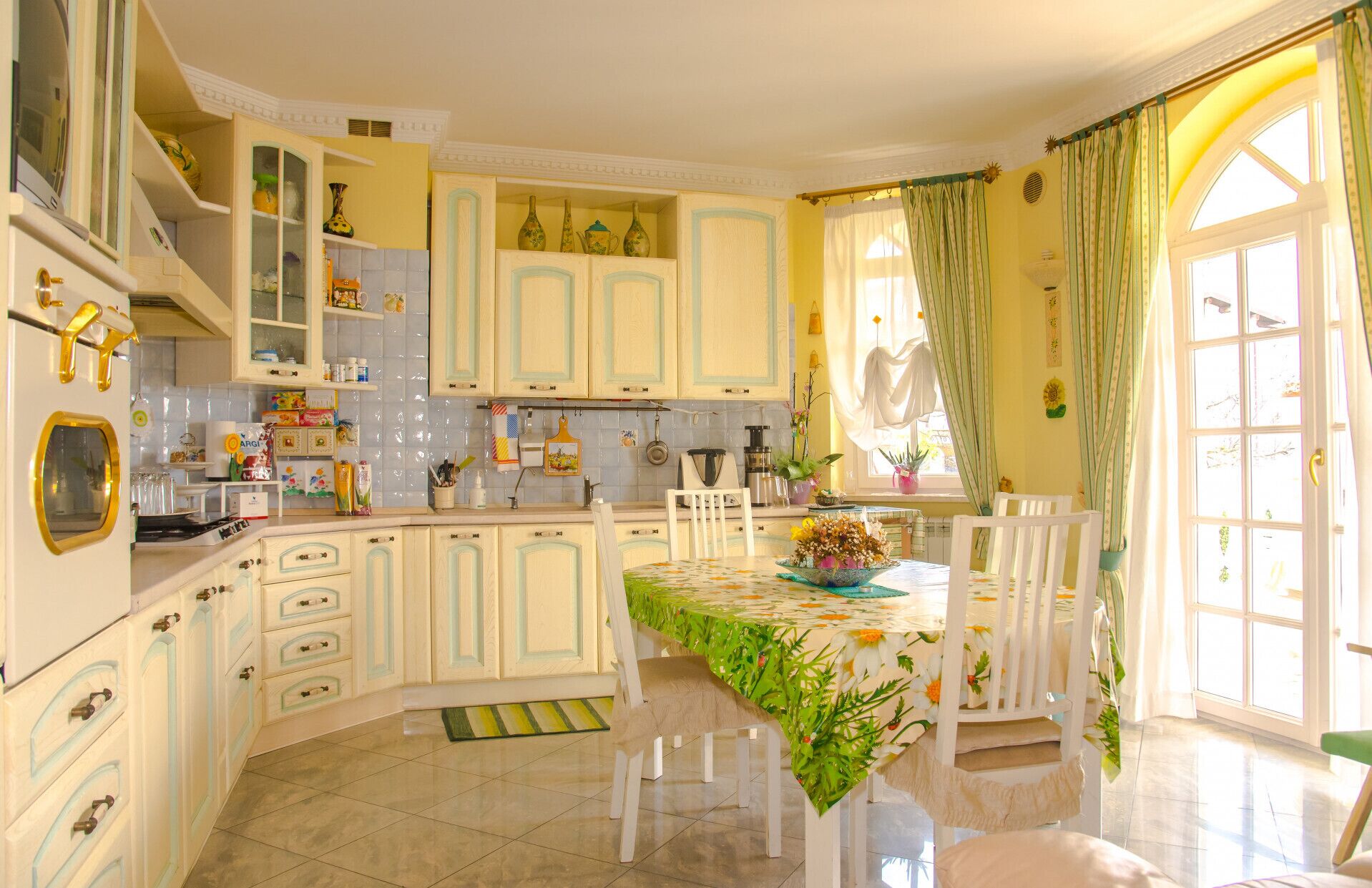 A kitchen with white cabinets, a dining table with a floral tablecloth, and a window with curtains.
