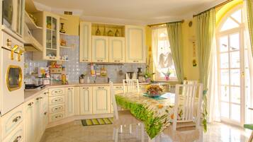 Une cuisine avec des meubles blancs, une table à manger recouverte d’une nappe à motifs floraux et une fenêtre avec des rideaux.