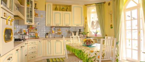 Une cuisine avec des meubles blancs, une table à manger recouverte d’une nappe à motifs floraux et une fenêtre avec des rideaux.