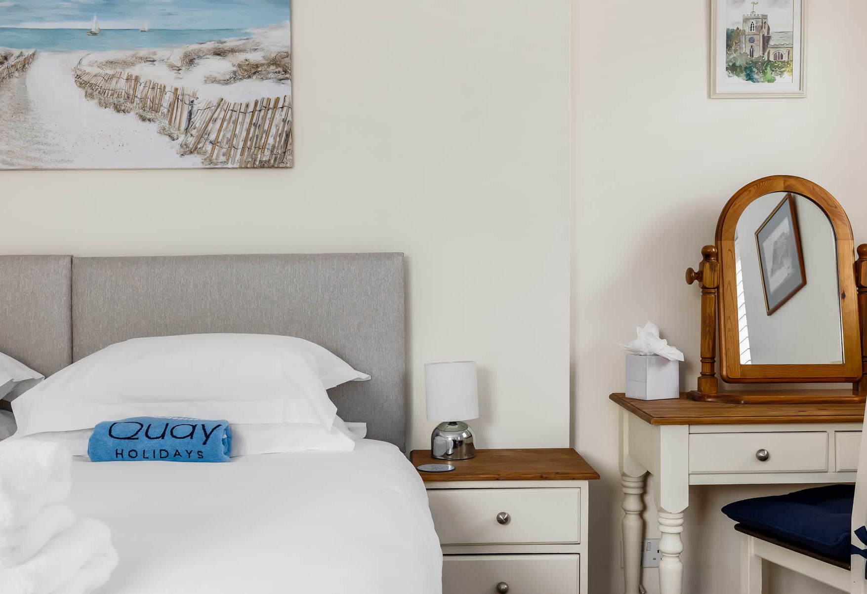 Habitación de hotel con una cama, una mesita de noche con lámpara, un aparador, un espejo y un cuadro de una playa.